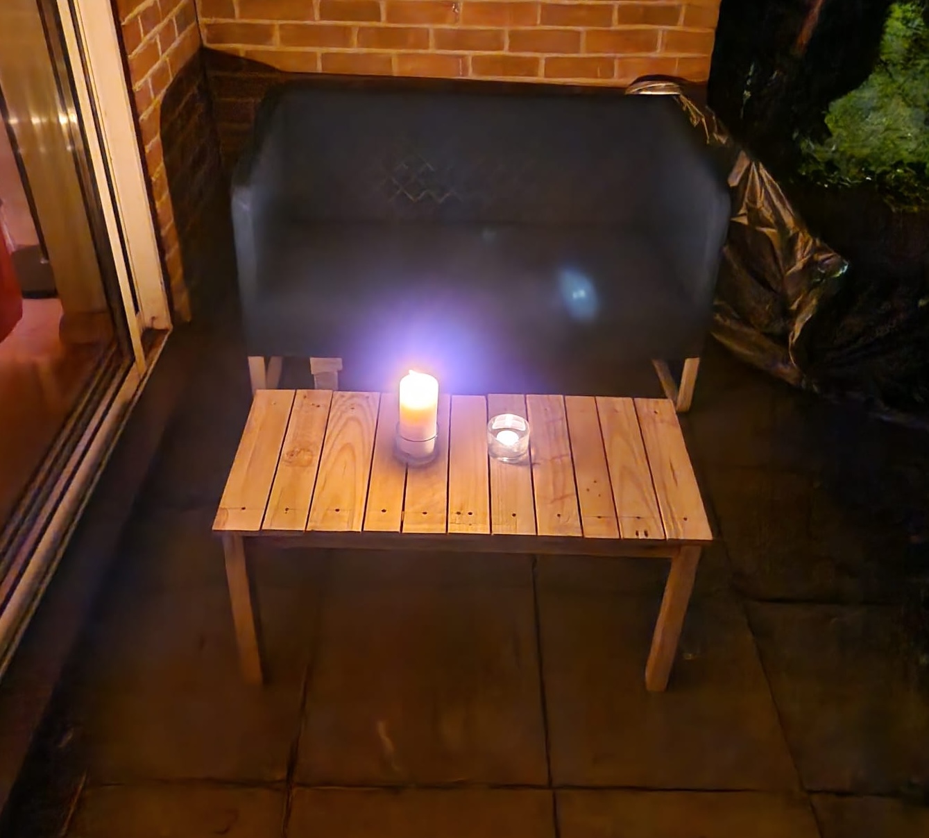 A small wooden table made of pallet wood with a candle in front of a small blue outdoor sofa on a patio at night