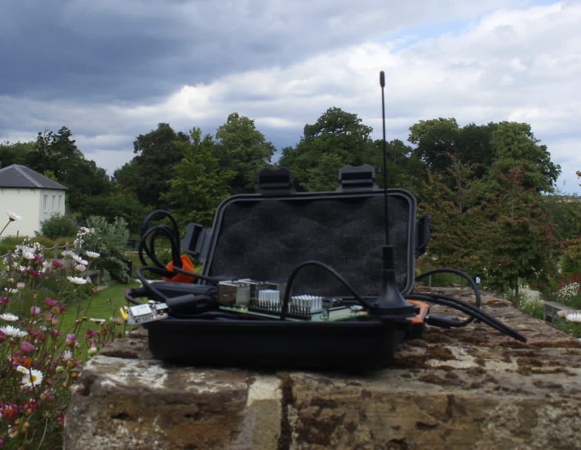 A small pelican case with a raspberry pi and an antenna on a wall in a park